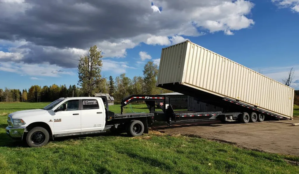 A 1-ton truck with a tilt-deck trailer delivering a 40' High Cube New (One-Trip) shipping container in an open field. The container is being smoothly offloaded, showcasing efficient and reliable delivery service in Canada.https://storecan.ca/+1 (800) 866-0208