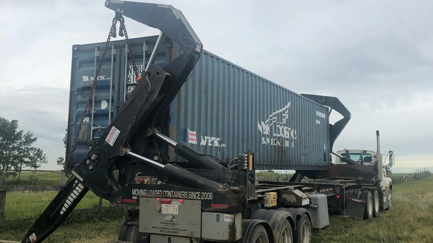A side loader truck delivering a 40' High Cube Used (Cargo-Worthy) shipping container in an open field. The container is being carefully placed on the ground, showcasing StoreCan’s efficient and reliable delivery services across Canada. Get high-quality shipping containers with fast delivery. Visit StoreCan.ca or call +1 (800) 866-0208.