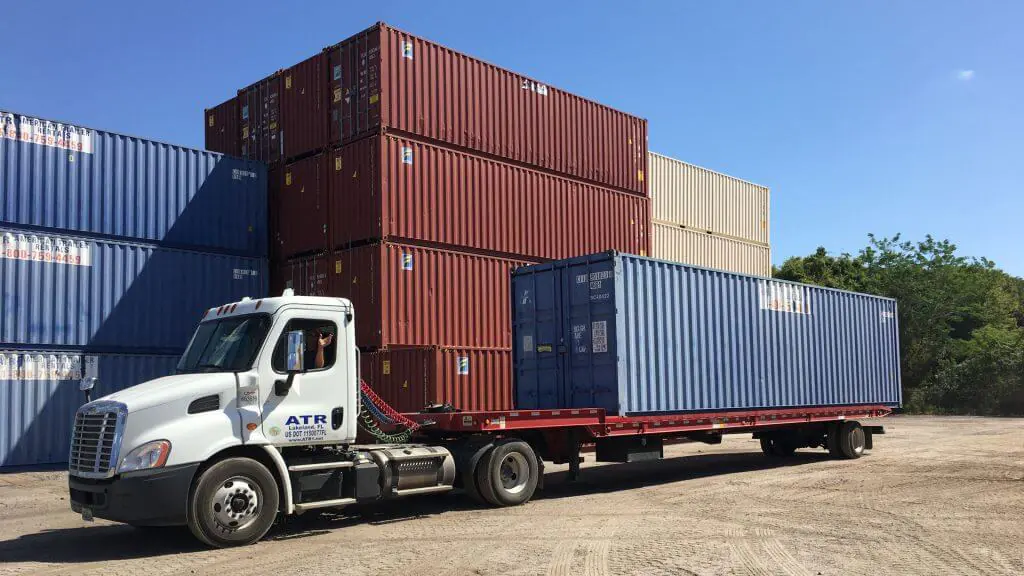 A semi-truck with a flatbed trailer picking up a 40' Used (Cargo-Worthy) shipping container at a StoreCan depot. The image highlights StoreCan’s reliable shipping container sales and transportation services across Canada. Get high-quality containers with fast delivery. Visit StoreCan.ca or call +1 (800) 866-0208.
